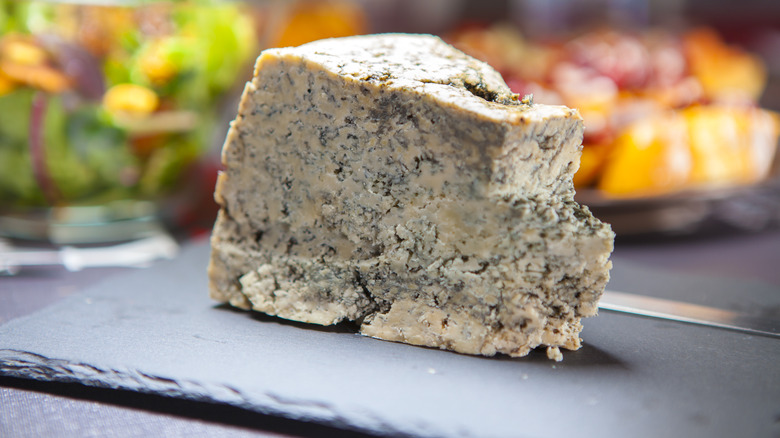 Close-up of a wedge of cabrales bleu cheese, made from cow, goat, and sheep's milk, from from Asturias, Spain on a stone platter with blurred fruits and vegetables in the background.