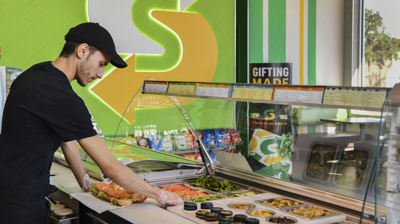 A Subway restaurant employee prepares a sandwich behind the counter.