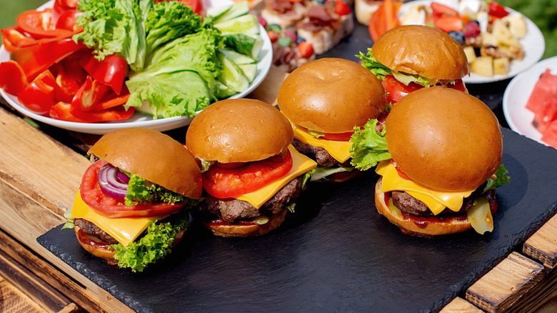 Several burgers in a cookout or picnic setting with lettuce and tomato in a bowl nearby