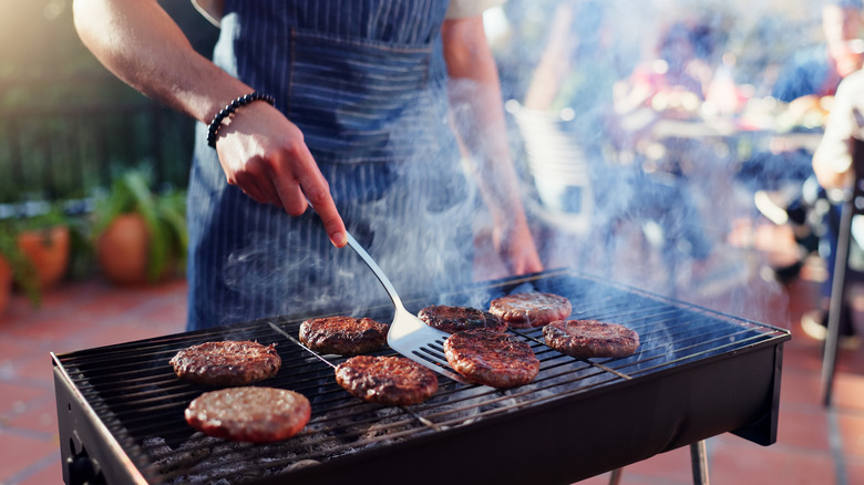 A person wearing an apron cooks burgers on a grill