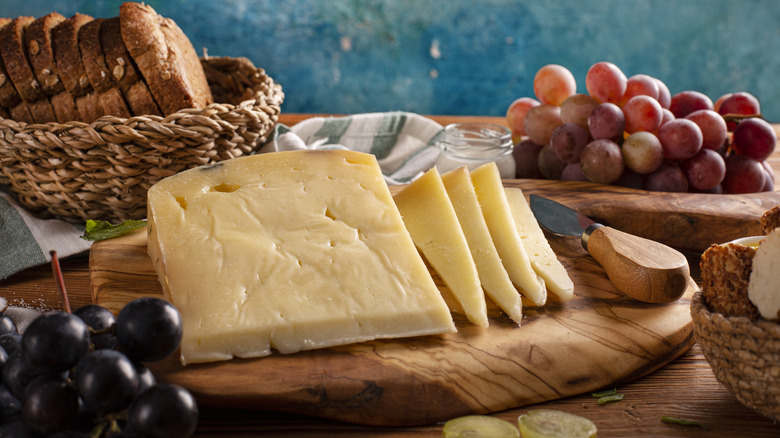 Gruyere cheese on wooden board surrounded by grapes and bread.