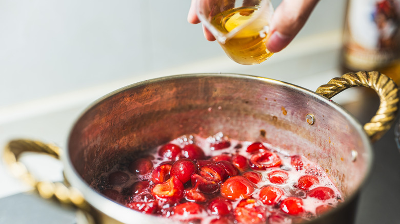 Person adding whiskey from a glass to a pot of cooking berries on a stove