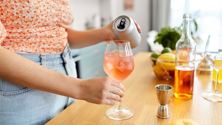 Woman pours a canned beverage into a wine glass with ice, with an unlabeled wine bottle and citrus around it