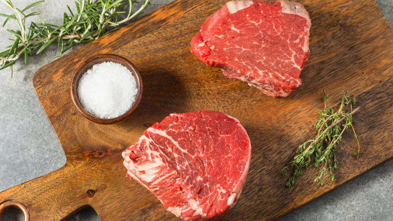 Two filet mignon steaks sit on a cutting board alongside herbs and a small bowl of salt.