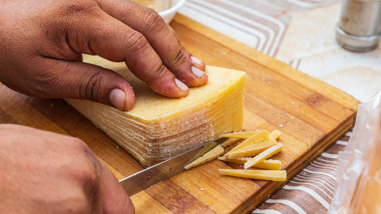 A block of cheese on a cutting board with someone using a knife to trim off the rind.