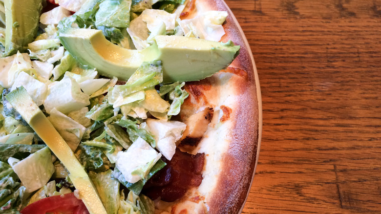 Pizza topped with avocado, lettuce, and cheese, served on a table.