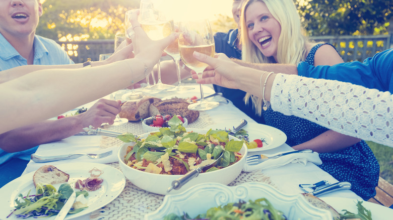 People sitting around a picnic table clinking glasses of white wine over a bowl of salad and bread