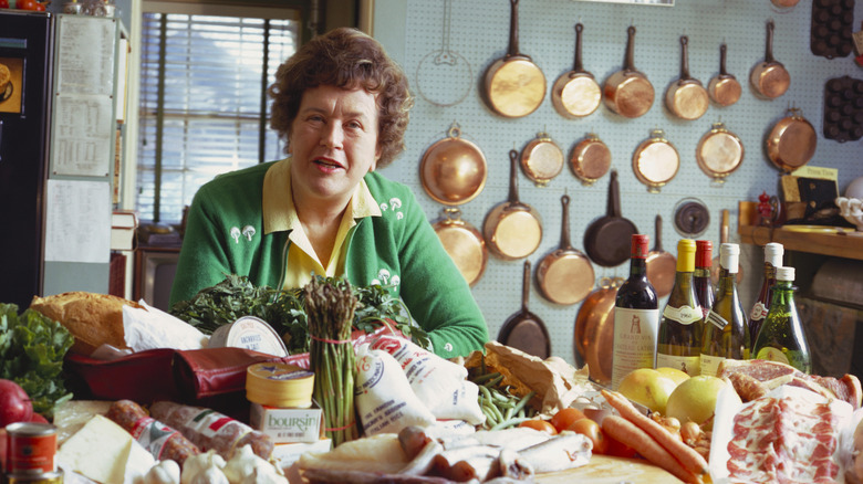 Julia Child in the kitchen surrounded by copper pots and ingredients like meat and produce.