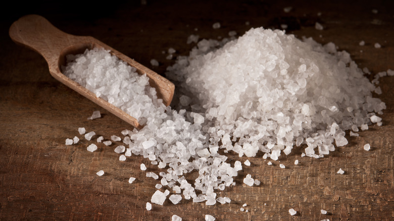 Chunks of salt in a pile on a wooden tabletop with a wooden scoop full of salt.