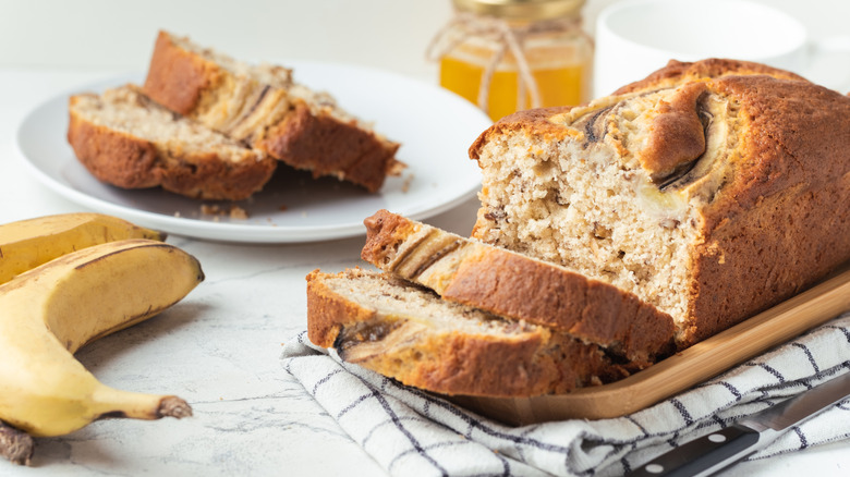 A lovely loaf of banana bread on a tray with slices on a plate in the background and two bananas on the lefthand side.