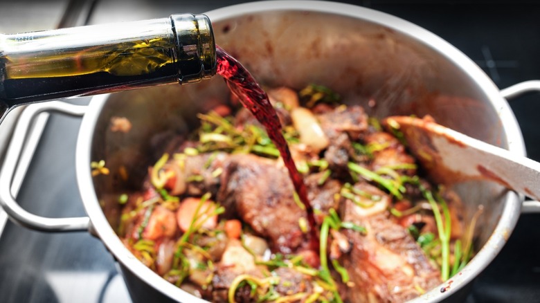 Red wine pouring from a bottle into an herbaceous pot of seared meat