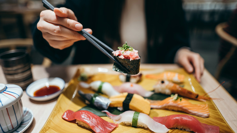 A well dressed person holding maki with black, textured chopsticks above a plate of nigiri sushi