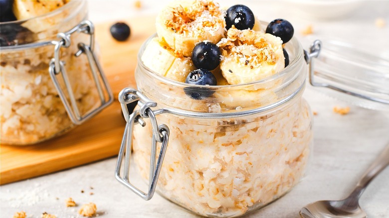 Open jars of cooked oatmeal with sliced banana, blueberries, and toasted coconut
