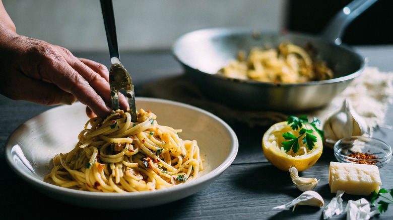 Pasta noodles covered in sauce being placed into a white bowl with garlic, cheese, spices, and a lemon rind on the tabletop next to it and a blurred image of a pan with pasta resting inside in the background