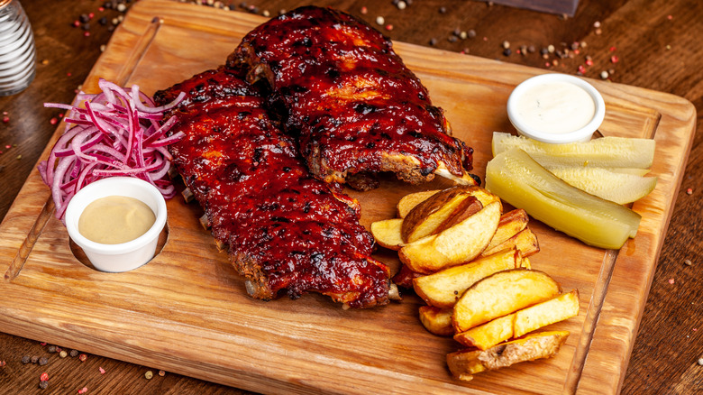 Slabs of cooked ribs on wood platter with pickles, potatoes and red onions